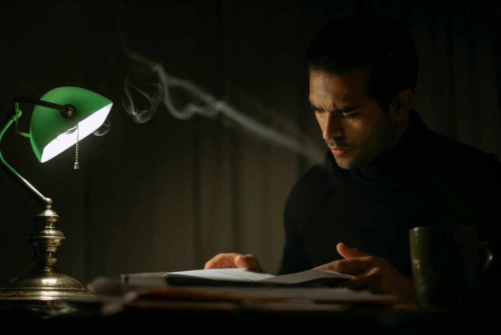 Pondering young male in black sweater reading book while sitting at desk with papers and green bankers lamp in dark home office
