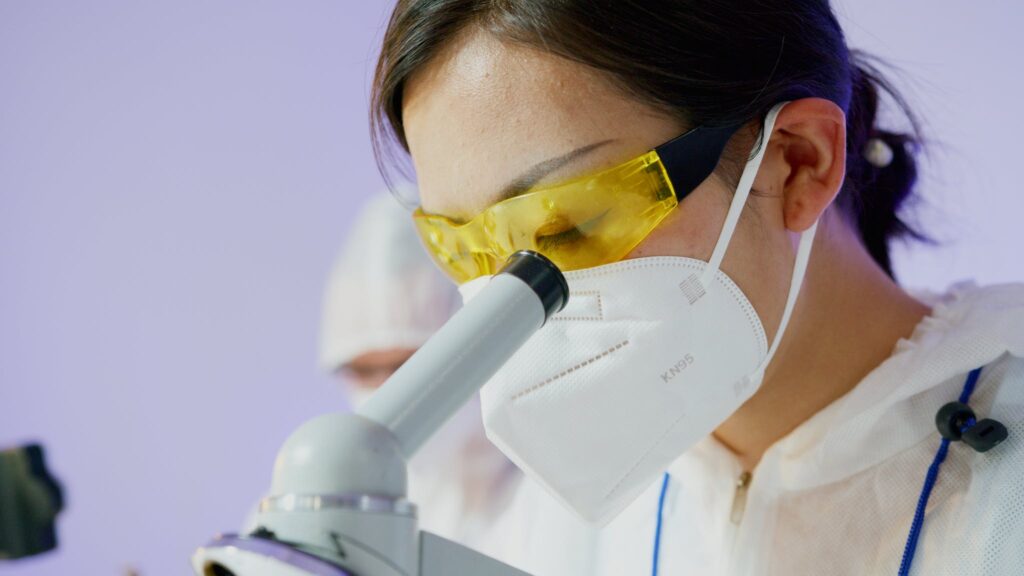 A Person Wearing Personal Protective Equipment Looking Through a Microscope