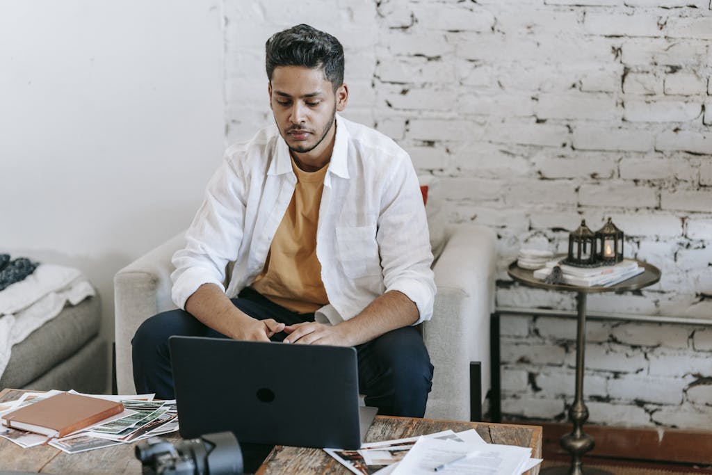 Creative young Hispanic male photographer sitting at table with photo camera and printed photos and working on laptop in modern loft style workplace at home